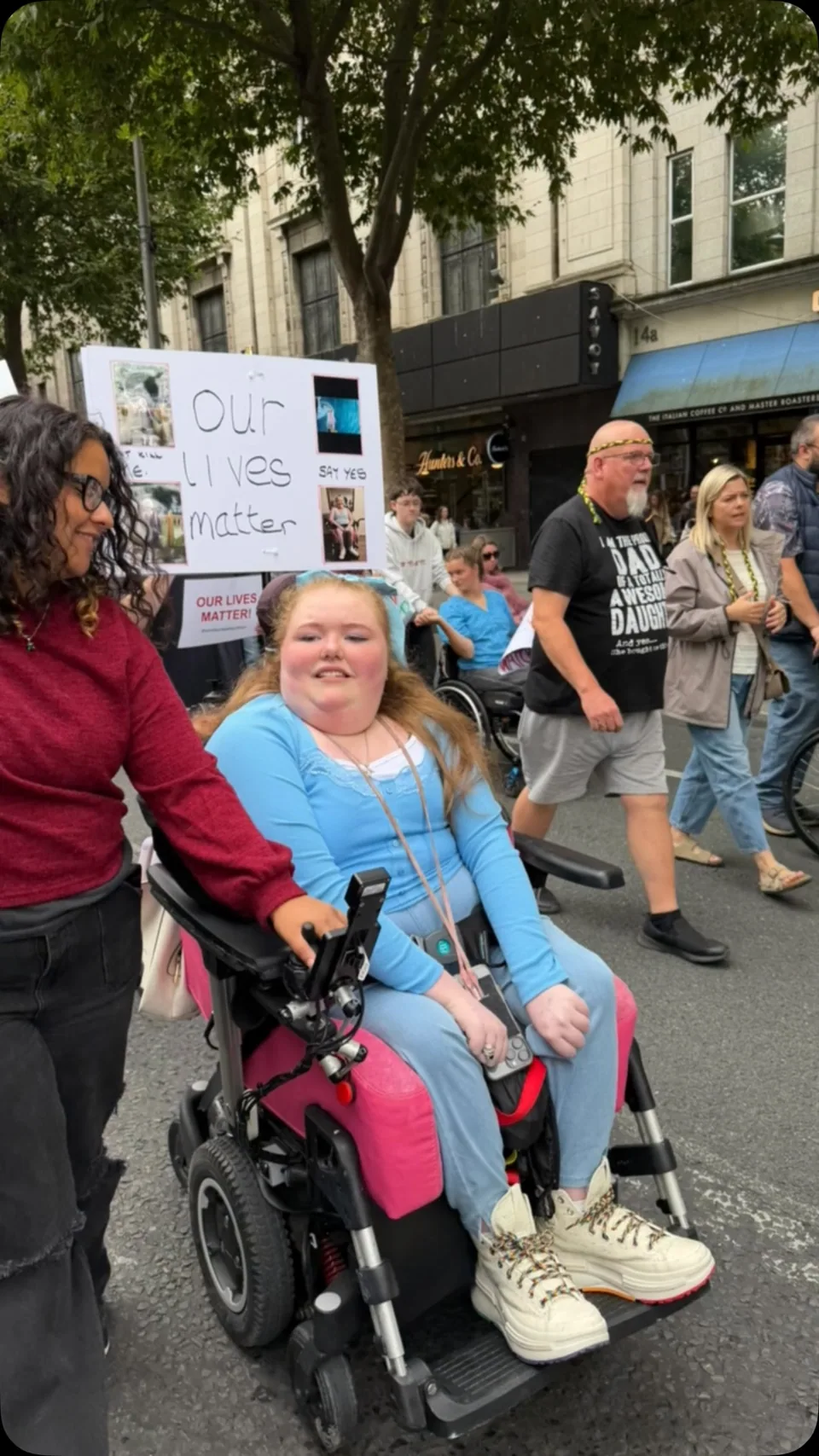 A person in a pink wheelchair at a protest holding a sign reading 'Our lives matter'